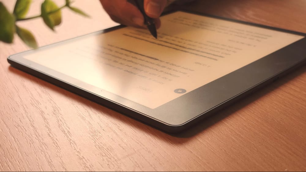 A close-up shot of a hand using a stylus to write on the screen of a Kindle Scribe. The writing style appears to be cursive. The context suggests note-taking, digital art, or form completion.